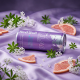 Cannette Red Bull Lilac Edition posée sur un tissu satiné violet avec fleurs de woodruff et quartiers de pamplemousse rose.