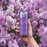 Main féminine tenant une canette Red Bull Lilac Edition devant un arbuste de lilas violet dans une ambiance printanière.